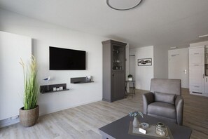 A modern living room with a flat-screen TV, a grey armchair, a coffee table, and a cabinet with glassware. - Holiday apartment with 1 bedroom (Cuxhaven)