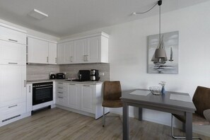 A modern kitchen with white cabinets, a built-in oven, a coffee maker, and a dining area with a table and chairs. - Holiday apartment with 1 bedroom (Cuxhaven)