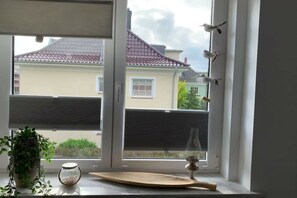 A window with a view of a house, a plant, a glass jar, and a wooden board. - Apartment by the sea (Travemünde)