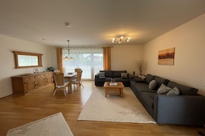 A living room with a sofa, a dining table with chairs, a kitchen area with cabinets, and a large window with curtains. - Strandpalais Apt. C1 (Cuxhaven)
