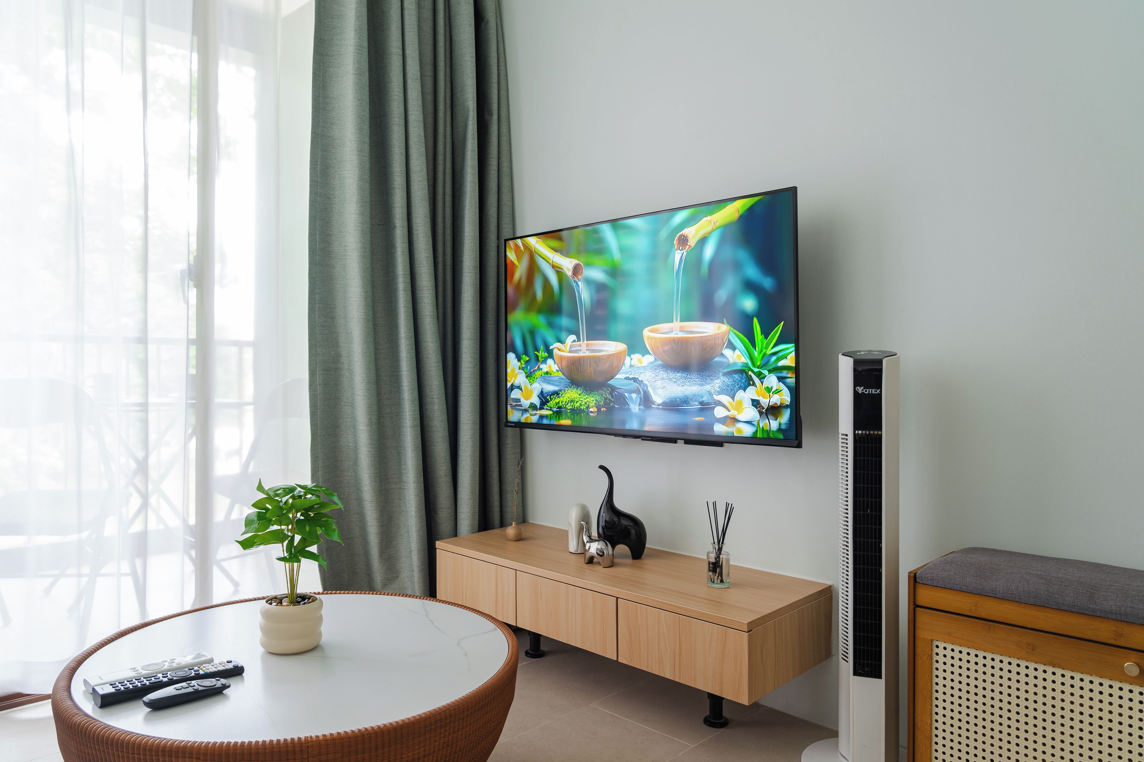 A modern living room with a flat-screen TV mounted on the wall, a round wooden table with a potted plant, a wooden cabinet with decorative items, and a standing heater.
