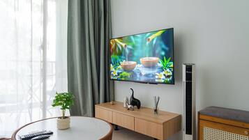 A modern living room with a flat-screen TV mounted on the wall, a round wooden table with a potted plant, a wooden cabinet with decorative items, and a standing heater.