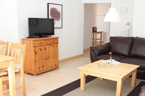 A living room with a TV on a wooden cabinet, a brown leather sofa, a wooden coffee table with decorative items, and a dining area with a wooden table and chairs. - in the forester's lodge on the Brocken mountain - 2 bedrooms (Ilsenburg)