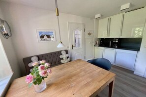 Uma sala de jantar com uma mesa de madeira, um vaso de flores, um relógio na parede e uma cozinha ao fundo.