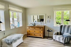 Quarto com cômoda de madeira, poltrona listrada, banco com manta de pele, espelho e janela com vista para área verde.