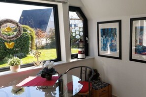 A dining area with a glass table, red cushion, and a view of a garden through a window. - Holiday home with 2 bedrooms (Butjadingen - Burhave)