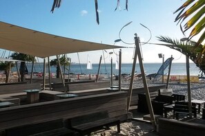 Ein Sitzbereich am Strand mit Holzbänken und Tischen, ein Segelboot im Hintergrund und freie Sicht auf das Meer.