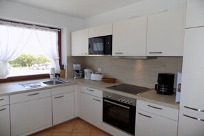 A compact kitchen with white cabinets, a built-in oven, a coffee maker, and a window with curtains. - House Oland Holiday Apartment 7 (Wenningstedt)