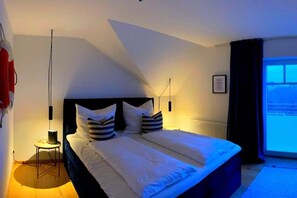 Un dormitori amb un llit gran, una tauleta de nit, una làmpada penjant, una finestra amb cortines i un radiador.