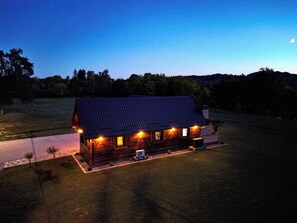 Una cabaña de madera con techo metálico, rodeada por un campo cubierto de hierba y árboles, bajo un cielo crepuscular.