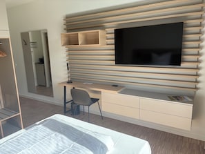 Quarto moderno com cama, escrivaninha com cadeira, TV de tela plana fixada em uma parede de painéis de madeira e uma pequena cozinha compacta ao fundo.