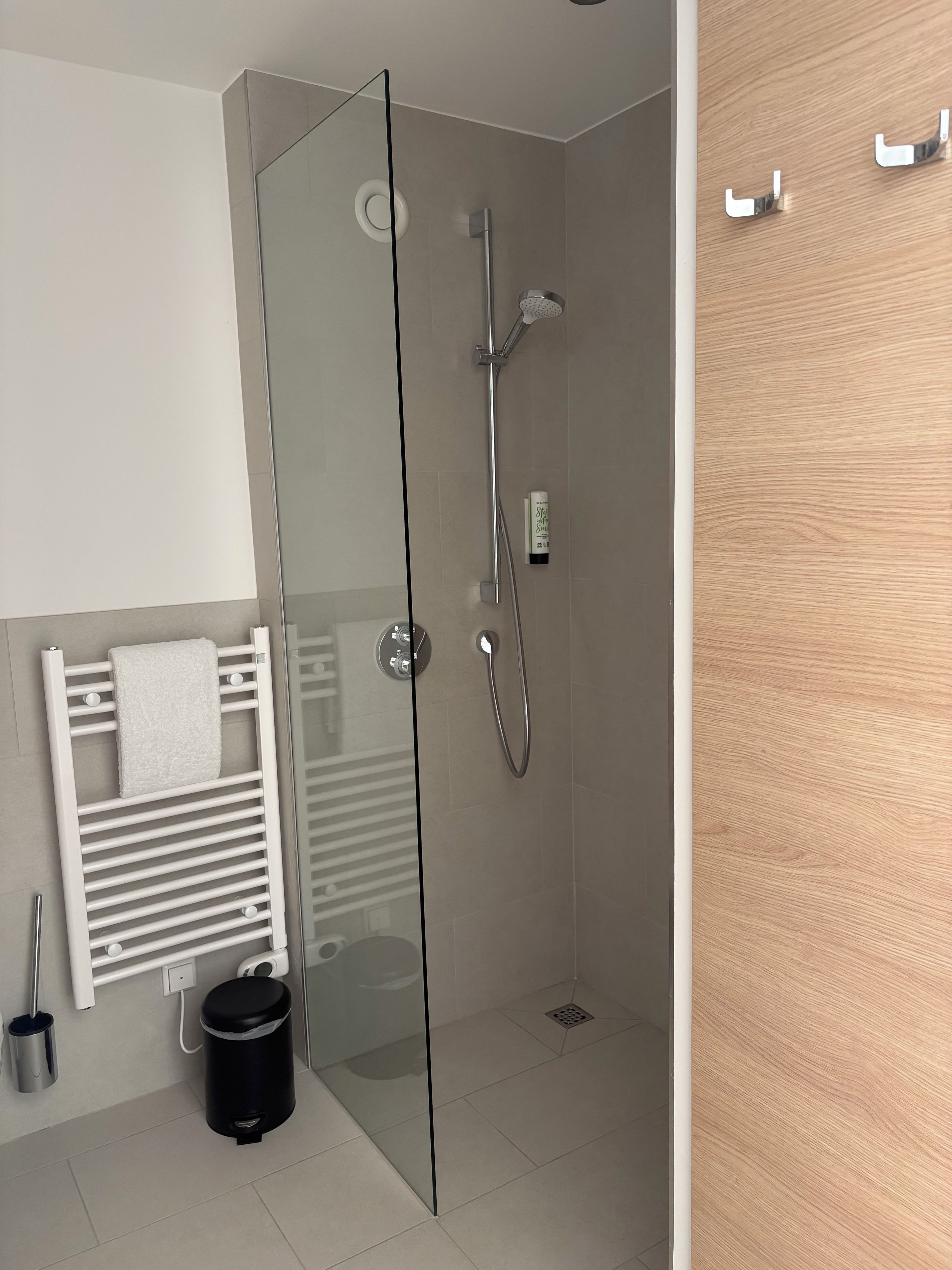 A modern bathroom with a glass shower enclosure, a white towel rack, and a black trash bin.