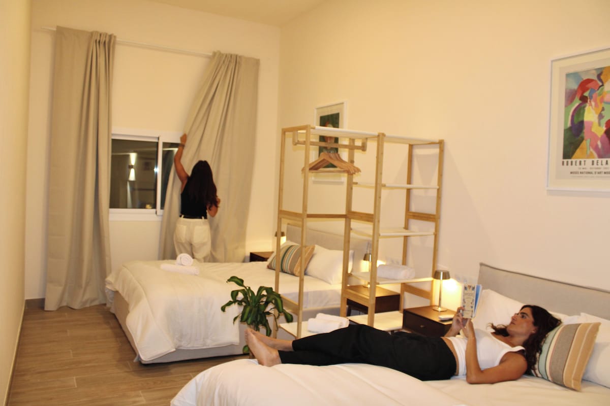 A hotel room with two beds, a wooden bunk bed, a window with curtains, a bedside table with a lamp, and a poster on the wall.