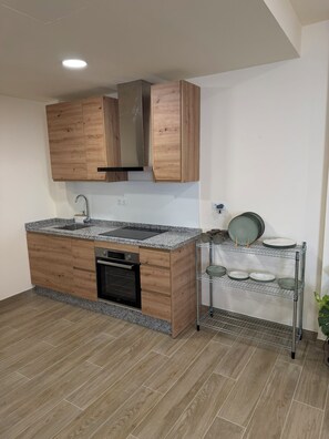 A kitchen with wooden cabinets, a stainless steel range hood, a built-in oven, a granite countertop, and a wire shelf holding plates and cups. - Lemon Tree Hostel (Málaga)