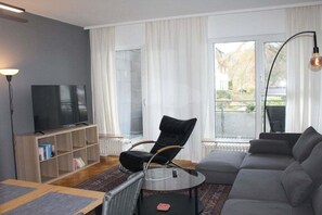 A modern living room with a grey sofa, a black chair, a glass coffee table, a flat-screen TV on a wooden stand, and a large window with curtains. - Tiny Atelier Bad Harzburg (Bad Harzburg)