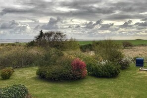 Eine Landschaft mit einem grasbewachsen Feld, dichter Strauchvegetation und einem fernen Blick auf das Meer unter einem bewölkten Himmel.