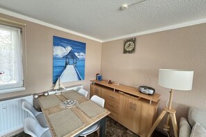 Ein Zimmer mit einem Esstisch, Stühlen, einer Uhr, einer Lampe und einem Gemälde eines Piers.