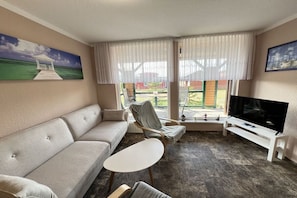 Sala de estar com sofá, poltrona, mesa pequena, televisão e janelas amplas com cortinas.