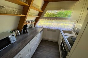 A compact kitchen with a built-in oven, a microwave, and a coffee maker. There is a window with a lace curtain, and shelves above the counter holding glassware. - Holiday home in Damp with barbecue and Wi-Fi (Damp)