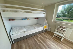 Een slaapkamer met een stapelbed, een raam, een klein bureau en een opbergbankje.