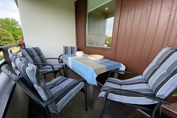 Een overdekt terras met rieten stoelen, een tafel, een raam en uitzicht op de omgeving.