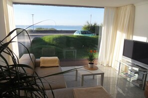 Uma sala de estar moderna com um sofá, uma mesa de centro, uma televisão e vista para o mar através de uma grande janela.