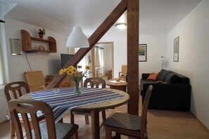 Een eethoek met een ronde tafel, stoelen en een vaas met zonnebloemen. Achterin staat een zwarte bank en een lamp.