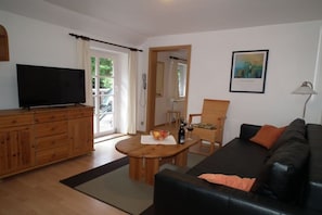 Ein Wohnzimmer mit Fernseher, einem Holzschrank, einem schwarzen Ledersofa, einem Esstisch mit Flasche und Gläsern sowie einem gerahmten Bild an der Wand.