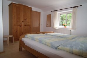 Ein Schlafzimmer mit einem hölzernen Bett, einem kleinen Nachttisch, einem Kleiderschrank, einem Fenster mit Vorhängen und einer Heizung.