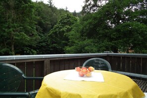 A table with a yellow tablecloth, a bowl of fruit, and a white napkin on a balcony with a wooden railing, surrounded by dense green foliage. - in the holiday home Nexö - 1 bedroom (Ilsenburg)