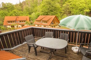 Eine Holzterrasse mit einem runden Tisch und Stühlen, umgeben von einem Holzzaun, mit Blick auf ein bewaldetes Gebiet und Häuser.