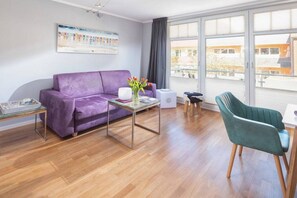 A living room with a purple sofa, a teal armchair, a glass coffee table, and a white speaker. - Holiday apartment with 1 bedroom (Norderney)