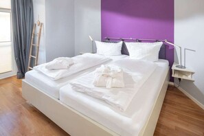 A double bed with white linens and two towels, a bedside table with a lamp, a wooden ladder, and a purple accent wall. - Holiday apartment with 1 bedroom (Norderney)