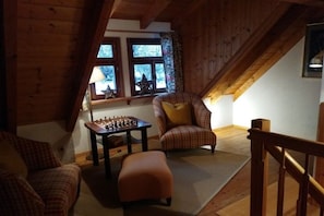 Ein gemütliches Zimmer mit Holzbalkendecke, einem Schachtisch, einem Karo-Sessel und einer Treppe.