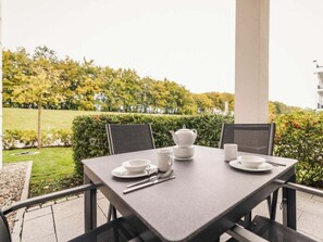 Ein Tisch für zwei Personen gedeckt mit Teekanne, Tassen und Besteck, mit Blick auf einen Garten mit Bäumen und einem Gebäude im Hintergrund.