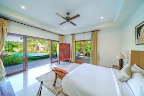Quarto com cama, mesa de cabeceira, luminária, ventilador de teto e vista para a piscina e jardim.