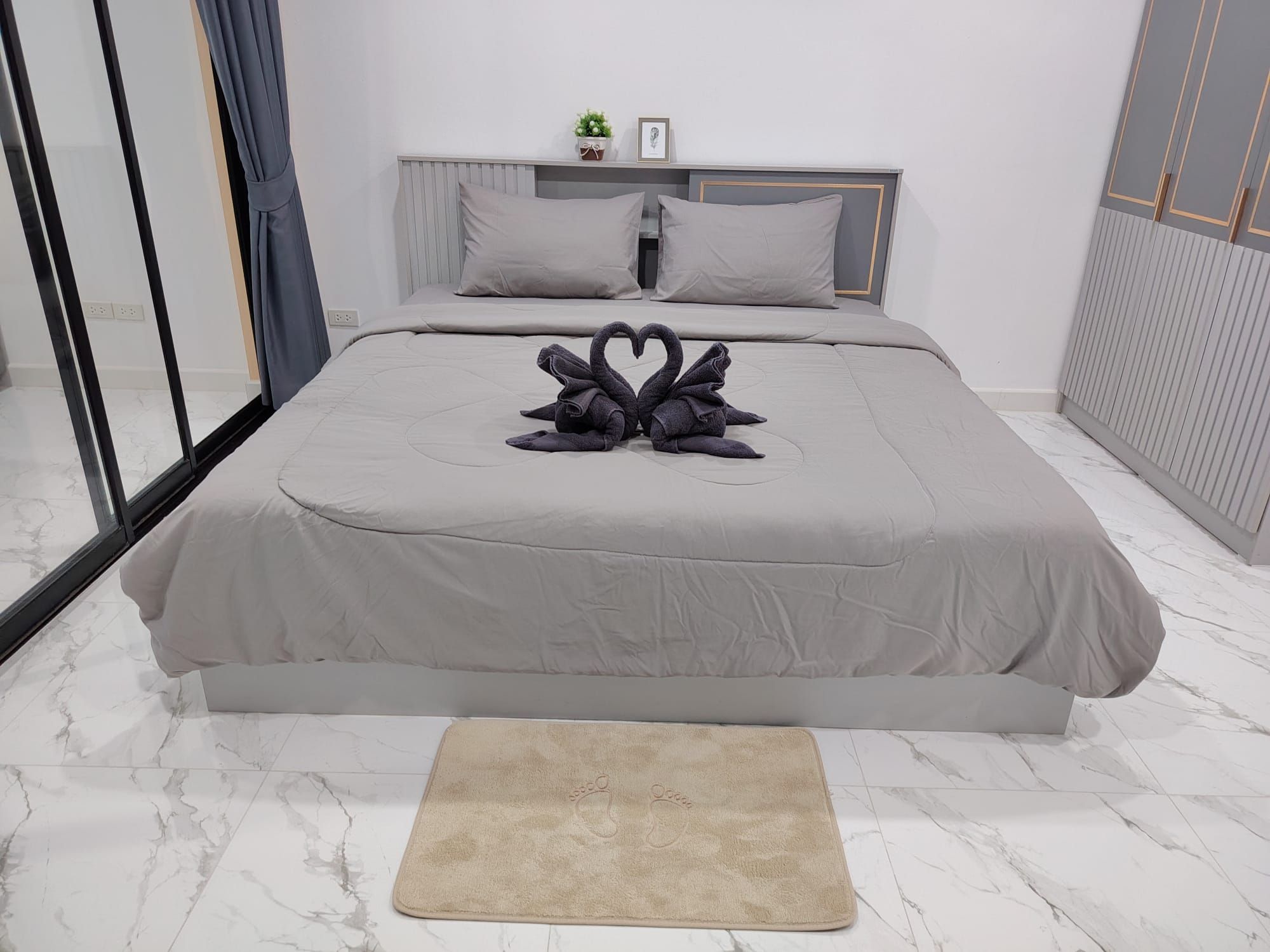 A modern bedroom with a large bed, a bedside table, a nightstand, a window with curtains, and a small potted plant.