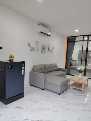 A modern living room with a grey sectional sofa, a small black refrigerator, a wooden coffee table, and a potted plant.