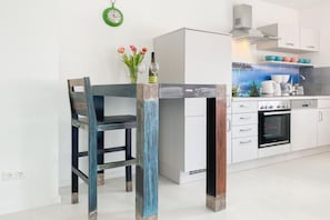 A modern kitchen with white cabinets, a built-in oven, and a dining area with a wooden chair and table. - Holiday home with 2 bedrooms (Timmendorfer Strand)
