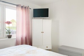A bedroom with a bed, a TV on a cabinet, a window with curtains, and a potted plant. - Holiday home with 2 bedrooms (Timmendorfer Strand)