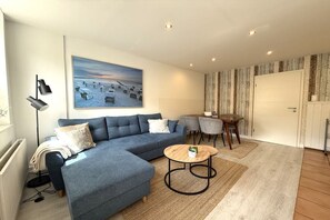 A modern living room with a sectional sofa, a round coffee table, and a painting of a beach scene. - Holiday apartment with 1 bedroom (Travemünde)