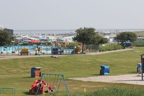 A grassy area with a playground, including swings and a slide, and a pool area with umbrellas and lounge chairs. - Holiday apartment in Nesse with swimming pool (Nesse)
