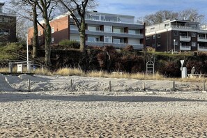 Ein mehrstöckiges Wohngebäude mit flachem Dach, einem eingezäunten Parkplatz und einem sandigen Vordergrund.