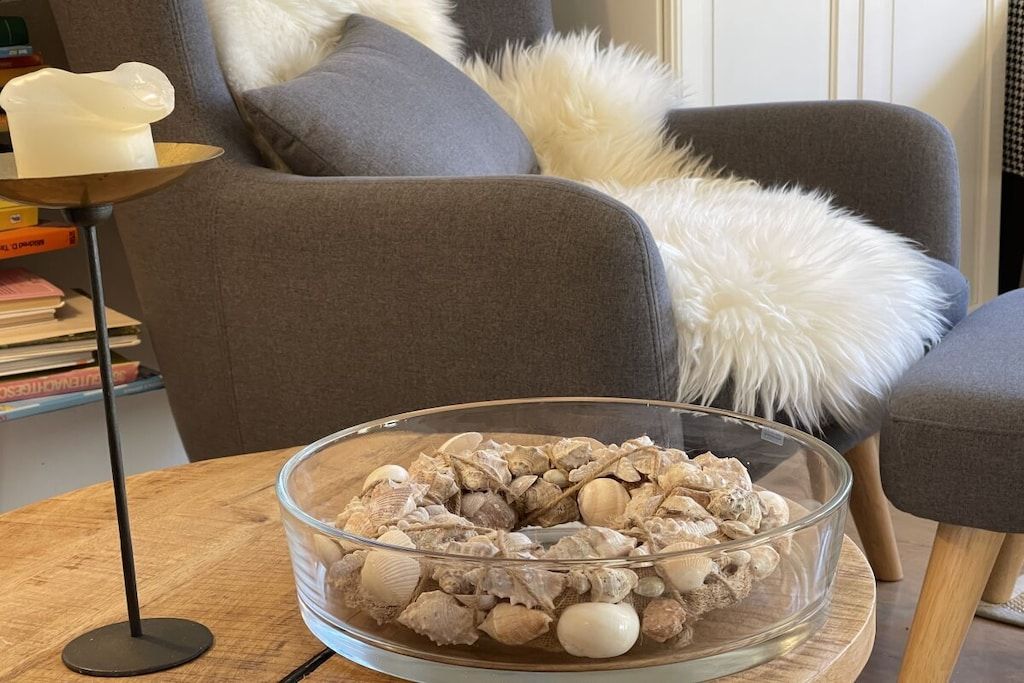 A clear glass bowl filled with various seashells on a wooden table, with a grey sofa and a white fluffy pillow in the background.