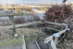 Une clôture en bois avec un panneau, un petit buisson et une vue sur un paysage où l’on aperçoit des personnes au loin.