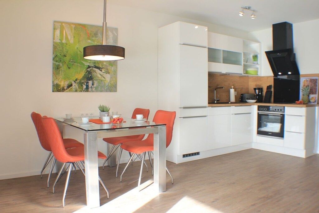 A modern kitchen with white cabinets, a dining table with orange chairs, and a hanging pendant light.