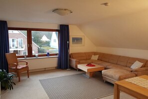 A cozy living room with a sectional sofa, a small coffee table, a chair, and a view of a house through a large window. - Holiday apartment with 2 bedrooms (Scharbeutz)