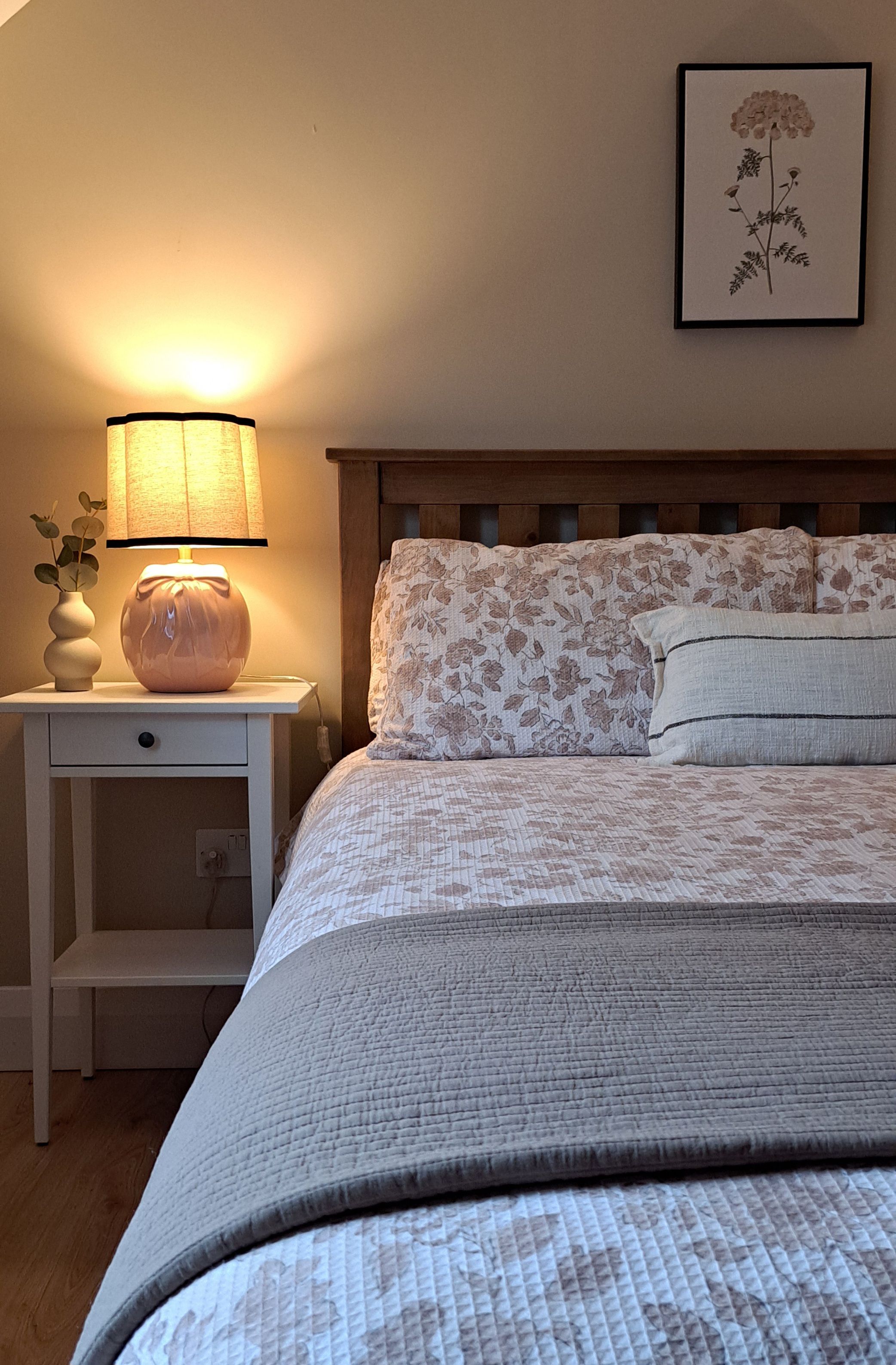 A bedroom with a bed, a nightstand with a lamp, a vase with a plant, and a framed picture on the wall.