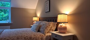 A bedroom with a bed, two bedside lamps, a window with a view of greenery, and a framed picture on the wall.