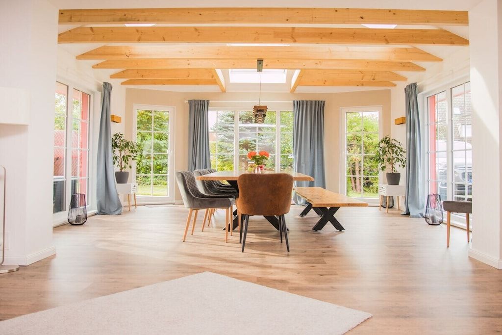A spacious room with a wooden ceiling, large windows with curtains, a dining table with chairs, and potted plants.